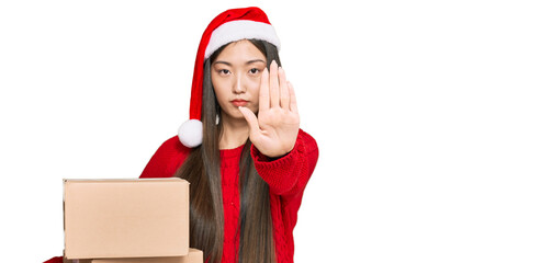 Canvas Print - Young chinese woman wearing christmas hat holding delivery package with open hand doing stop sign with serious and confident expression, defense gesture