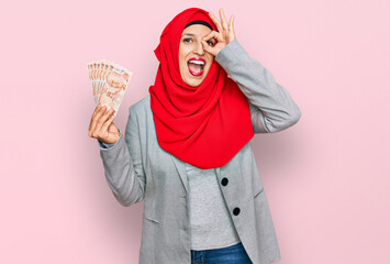 Sticker - Beautiful hispanic woman wearing islamic hijab holding turkish lira banknotes smiling happy doing ok sign with hand on eye looking through fingers