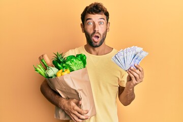 Poster - Handsome man with beard holding groceries and 20 swedish krona banknotes afraid and shocked with surprise and amazed expression, fear and excited face.