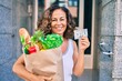 © Krakenimages.com - Middle age hispanic woman smiling happy holding a grocery shopping bag full of groceries and american 20 dollar at the city.