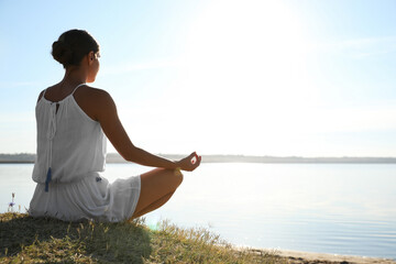 Wall Mural - Young woman meditating near river at sunset, space for text. Nature healing power