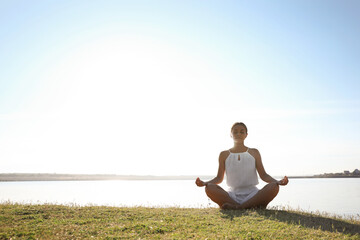 Wall Mural - Young woman meditating near river at sunset, space for text. Nature healing power