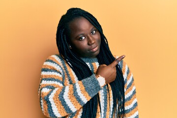 Sticker - Young black woman with braids wearing casual winter sweater pointing with hand finger to the side showing advertisement, serious and calm face