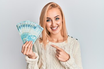 Wall Mural - Beautiful caucasian blonde woman holding 100 brazilian real banknotes smiling happy pointing with hand and finger