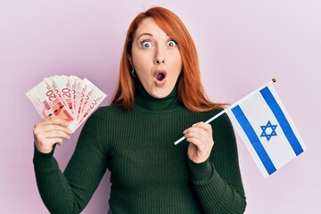 Poster - Beautiful redhead woman holding 20 shekels banknotes and israel flag afraid and shocked with surprise and amazed expression, fear and excited face.