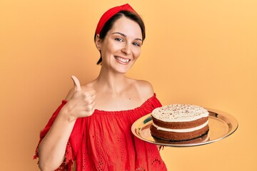 Young brunette woman with short hair holding carrot cake smiling happy and positive, thumb up doing excellent and approval sign