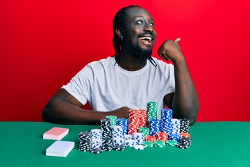 Poster - Handsome young black man sitting on the table with poker chips and cards smiling with happy face looking and pointing to the side with thumb up.