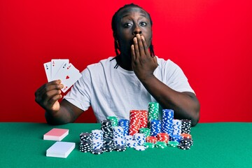 Wall Mural - Handsome young black man playing poker holding cards covering mouth with hand, shocked and afraid for mistake. surprised expression