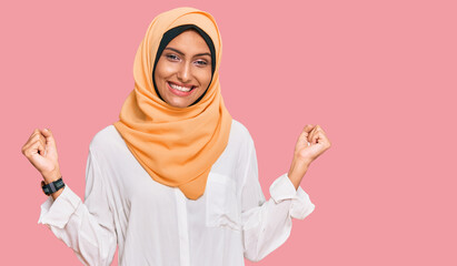 Poster - Young brunette woman wearing traditional islamic hijab scarf very happy and excited doing winner gesture with arms raised, smiling and screaming for success. celebration concept.