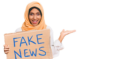 Sticker - Young brunette arab woman holding fake news banner celebrating victory with happy smile and winner expression with raised hands