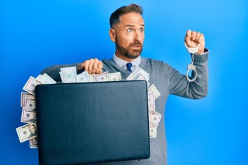 Poster - Handsome middle age man holding briefcase full of dollars and handcuffs in shock face, looking skeptical and sarcastic, surprised with open mouth