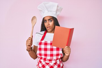 Wall Mural - Young african american woman with braids wearing professional baker apron reading cooking recipe book clueless and confused expression. doubt concept.