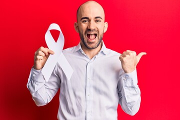 Sticker - Young handsome bald man holding white cancer ribbon pointing thumb up to the side smiling happy with open mouth