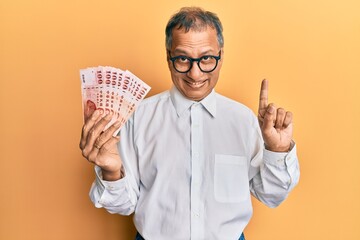 Sticker - Middle age indian man holding 100 new taiwan dollars banknotes smiling with an idea or question pointing finger with happy face, number one