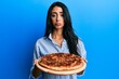 Beautiful hispanic woman holding tasty pepperoni pizza depressed and worry for distress, crying angry and afraid. sad expression.