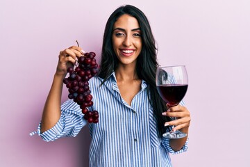 Wall Mural - Beautiful hispanic woman holding branch of fresh grapes and red wine smiling and laughing hard out loud because funny crazy joke.