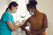 Young African American Woman Getting Flu Shot During Seasonal Vaccination Campaign. Doctor Or Nurse In Medical Face Mask Cleans Skin On Patient's Arm Before Injecting Modern Covid 19 Antiviral Vaccine