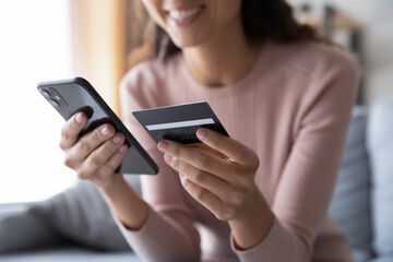 Close up smiling millennial woman holding smartphone and banking credit card, involved in online mobile shopping at home, happy female shopper purchasing goods or services in internet store.