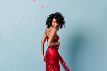 excited curly girl posing under confetti. studio shot of carefree woman in red dress.