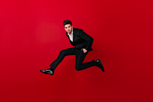 Good-looking Man In Stylish Suit Jumping On Red Background. Studio Shot Of Brunette Guy Having Fun.