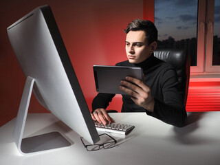 Poster - Young businessman works in his home office. Man at a remote job. Home office of a young businessman. Guy sits at a table next to a computer and holds a tablet in his hands. Gadgets for business.