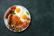 Full English Breakfast On A Plate With Fried Eggs, Sausages, Bacon, Beans, Toasts And Coffee On Dark Stone Background. With Copy Space. Top View