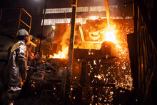 Foundry And Industrial Steel Production. Worker In Aluminized Protection Suit Standing By Furnace With Hot Molten Iron And Sparks Flying.