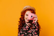 Positive blue-eyed redhead girl in floral print top happily makes photo on pink camera