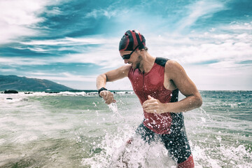 Triathlete swimmer looking at sport watch app using smartwatch during triathlon. Swimming man running out of ocean swim checking heart rate on smart watch. Professional athlete training for ironman.