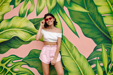 Gorgeous Young Woman In Shorts Standing In Front Of Green Graffiti. Outdoor Shot Of Girl In Pink Sunglasses.