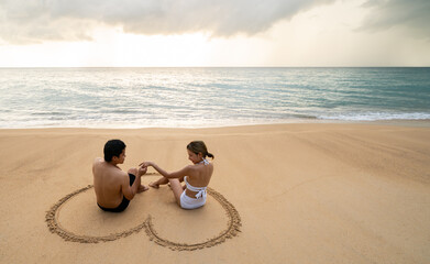 Couple sitting on the beach in drawing heart shape on sand. During sunset in the summer.