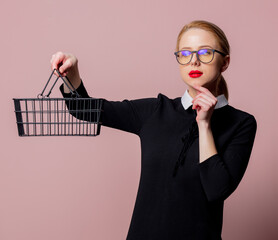 Wall Mural - Blonde woman black dress and glasses with shopping basket on pink background