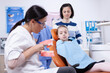© DC Studio - Ortodontist brushing artificial jaw educating child sitting on dental chair. Little girl and mother listening stomatolog talking about tooth hygine in dentistiry clinic holding jaw model.
