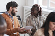 © Seventyfour - Multi-ethnic group of people in audience at business conference focus on bearded man talking to African-American woman while sitting on chairs