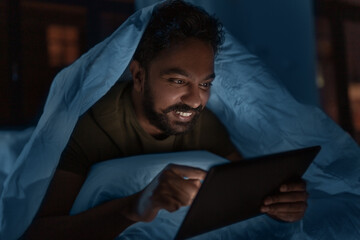 Poster - technology, internet, communication and people concept - young indian man with tablet pc computer lying in bed under blanket at home at night