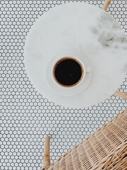 Wall Mural - Flat lay, top view of cup of coffee on white round marble table with rattan chair on vintage black and white mosaic. Minimal background with neutral colors.