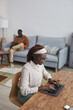 © Seventyfour - Vertical high angle portrait of young African-American woman using laptop at desk while working from home in cozy apartment interior