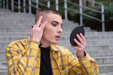 Portrait of a handsome young man, looking at his wrinkles in front of a mirror, wearing black eyeshadow, holding a makeup brush. Non binary androgynous guy.