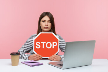 Wall Mural - Serious woman office worker holding stop symbol, warning with red traffic sign and looking angrily, sitting at workplace with laptop, home office. Indoor studio shot isolated on pink background