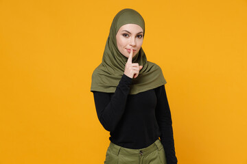 Wall Mural - Secret young arabian muslim woman in hijab black green clothes saying hush be quiet with finger on lips shhh gesture isolated on yellow background studio portrait. People religious lifestyle concept.