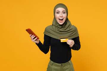 Wall Mural - Surprised young arabian muslim woman in hijab black green clothes using mobile cell phone hold credit bank card isolated on yellow color background studio portrait. People religious lifestyle concept.