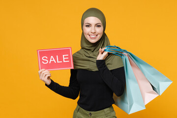 Wall Mural - Smiling young arabian muslim woman in hijab green black clothes hold package bags with purchases after shopping sign with SALE title isolated on yellow background. People religious lifestyle concept.