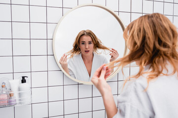 Wall Mural - displeased woman in bathrobe adjusting hair near mirror in bathroom