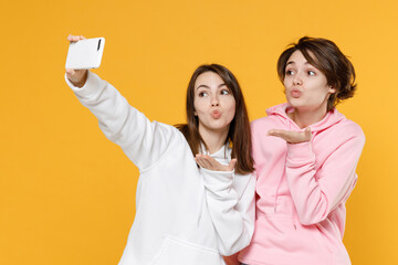 Wall Mural - Pretty two young women friends 20s wearing casual white pink hoodies standing doing selfie shot on mobile phone blowing sending air kiss isolated on bright yellow color background studio portrait.