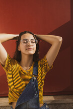 Portrait Of A Beautiful Caucasian Teenager Girl Wearing Transparent Trendy Glasses And Colorful Clothes With Her Hands Behind Her Head Smiling With Eyes Shut In Relief. Dark Red Background. 