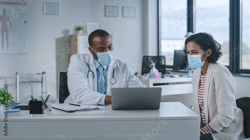 Family Doctor in Protective Mask is Reading Medical History of Female Patient and Speaking with Her During Consultation in a Health Clinic. Physician in Lab in Front of Computer in Hospital Office.