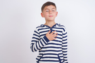 Satisfied smiling little cute boy kid wearing stripped t-shirt against, keeps hands on belly, being in good mood after eating delicious supper, demonstrates she is full. Pleasant feeling in stomach.