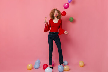 Wall Mural - Full-length shot of beautiful curly blonde woman in stylish jeans, red blouse holding glass of wine and having fun among balloons