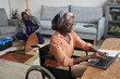 © Seventyfour - Portrait of modern African-American woman using wheelchair and working from home with handyman assembling furniture in background, service and assistance concept, copy space