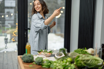 Wall Mural - Beautiful young woman dancing on the kitchen while cooking healthy green fresh salad. Vegetarian clean organic lifestyle. High quality photo